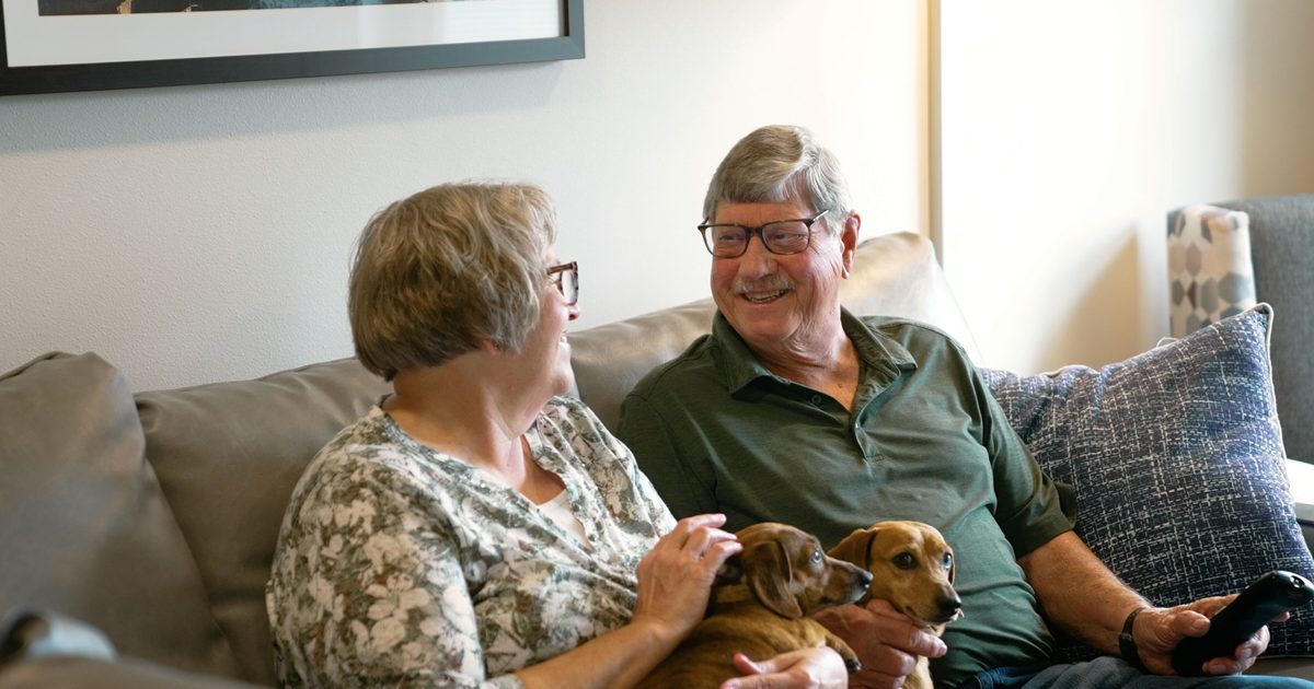 An older couple sitting on a couch with two dogs in their lap