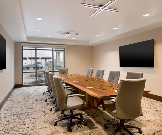 A boardroom containing a large table with chairs. There are TVs on either wall and a large window at the end of the room.